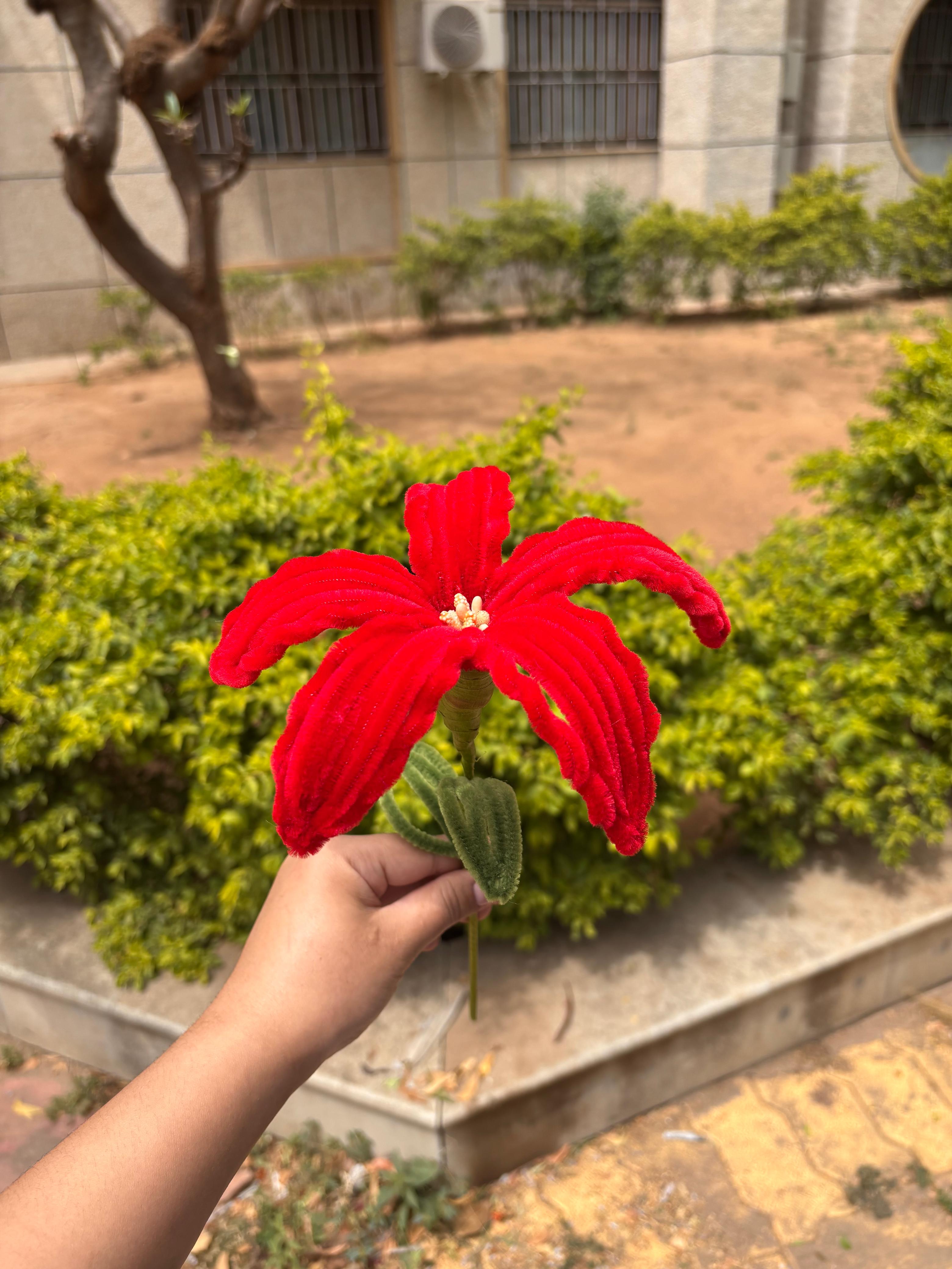 Red artificial flower held by a hand with a garden and building in the background